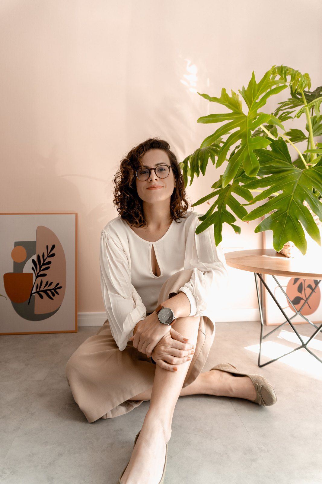 Renata sentada no chão ao lado de uma planta e de um quadro